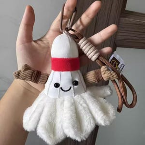 Pode incluir: Um chaveiro de volante de badminton de pelúcia com um rosto sorridente, penas brancas e uma faixa vermelha. O chaveiro tem um cordão marrom e uma pequena etiqueta.