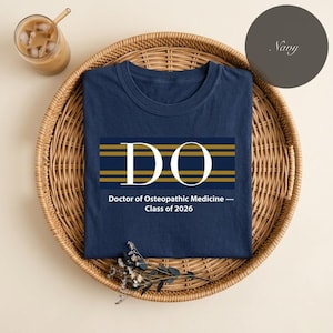 Op de afbeelding: Marineblauw T-shirt met een wit en goud ontwerp met de tekst "DO" boven de tekst "Doctor of Osteopathic Medicine - Class of 2026". Het shirt wordt gepresenteerd op een geweven dienblad met een glas ijskoffie.