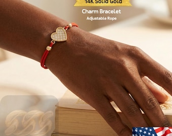 May include: A red adjustable rope charm bracelet with a heart-shaped pendant. The pendant is encrusted with small, clear stones and is framed in gold. The text "14K Solid Gold Charm Bracelet Adjustable Rope" is visible.