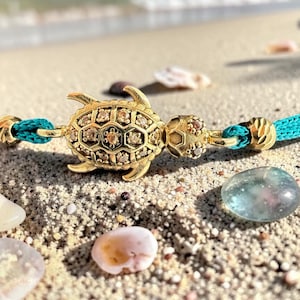 May include: A gold-colored turtle charm bracelet with a turquoise cord. The turtle is encrusted with small, sparkling stones. The bracelet is resting on a sandy surface with seashells and a smooth, blue-green stone.