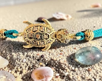 May include: A gold-colored turtle charm bracelet with a turquoise cord. The turtle is encrusted with small, sparkling stones. The bracelet is resting on a sandy surface with seashells and a smooth, blue-green stone.