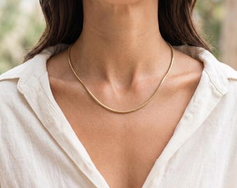 May include: A gold-toned snake chain necklace with a smooth, flexible, and rounded link design. The necklace rests against the collarbone, featuring a polished metallic finish that reflects light evenly across its continuous, uniform surface.
