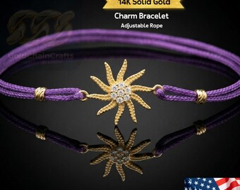 May include: A purple charm bracelet with a gold sunburst pendant. The pendant is encrusted with small, clear gemstones. The bracelet has an adjustable rope design and is labeled as 14K solid gold.