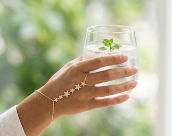 May include: A gold-toned hand chain bracelet featuring a series of five small, star-shaped charms encrusted with tiny clear stones. The delicate chain connects a wrist bracelet to a single ring, draping across the back of the hand.