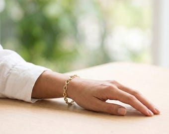 Puede incluir: Una pulsera de cadena dorada con eslabones ovalados entrelazados en la muñeca. La pulsera está hecha de metal dorado pulido y consta de una serie de eslabones uniformes y conectados que forman una banda circular flexible.