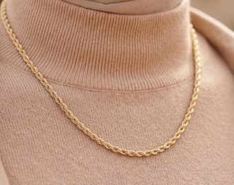 May include: A gold-toned rope chain necklace featuring a twisted, spiral link design. The chain rests against a beige ribbed turtleneck sweater, showcasing a uniform metallic finish and a continuous, interconnected circular pattern throughout the length.
