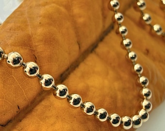 May include: Close-up of a gold ball chain necklace. The necklace is composed of small, round, polished gold beads linked together. The chain is resting on a textured, yellow-brown surface, creating a contrast between the shiny metal and the organic background.