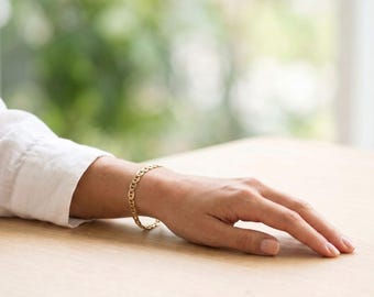 May include: A gold chain bracelet featuring a series of interlocking oval links. The bracelet is worn on a wrist, showcasing the polished metallic finish and the consistent, repeating pattern of the circular and oval-shaped metal links.