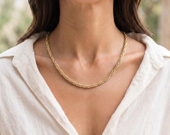 May include: A gold Byzantine chain necklace featuring a series of interlocking, woven metal links that create a rounded, textured appearance. The necklace rests against the neck, showcasing a polished, reflective finish in a warm yellow gold tone.