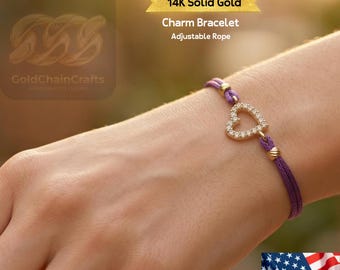 May include: A 14K solid gold charm bracelet with an adjustable purple rope. The bracelet features a heart-shaped charm adorned with small, sparkling stones. The text "14K Solid Gold Charm Bracelet Adjustable Rope" is visible.