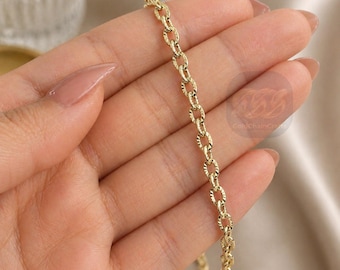 May include: Close-up of a gold-colored chain necklace with an oval link design. The chain is held in a hand with a neutral-toned manicure. The chain has a textured pattern on each link. The background is a soft, neutral color.