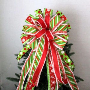 May include: A festive Christmas tree topper bow. The bow features red, green, and white ribbons with snowflake, polka dot, and striped patterns. The bow is made of multiple layers of ribbon and is designed to sit atop a Christmas tree.