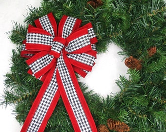 May include: A Christmas wreath with a large bow. The bow has a red border and a black and white houndstooth pattern. The wreath is made of green pine needles and has several pine cones.