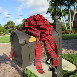 May include: A brown mailbox adorned with a large red and black buffalo plaid bow. The mailbox has a gold-colored address plate with the number 54 and the name "ROSS" visible. The scene is set outdoors with a blue sky and palm trees.
