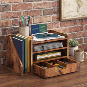 May include: A wooden desk organizer with multiple compartments and a drawer. The organizer holds notebooks, files, pens, and business cards. The text "YOUR NAME" is visible on a business card. The organizer is made of brown wood and sits on a wooden desk.