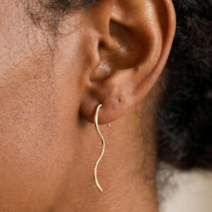 May include: A gold-toned earring featuring a thin, vertical, wavy wire design that hangs from the earlobe. The metal has a smooth, polished finish and follows a gentle, serpentine curve that extends downward from the point of attachment.