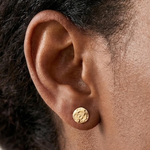 May include: A close-up of a gold-colored stud earring with a textured surface. The earring is round and appears to be made of metal. The earring is in the ear of a person with dark skin and dark hair.