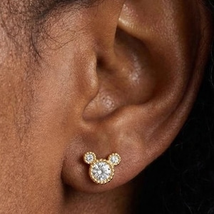 May include: Gold-toned stud earrings in a Mickey Mouse design. The center of the mouse's head features a round, clear gemstone surrounded by smaller clear stones. The ears are represented by two smaller, round clear stones.