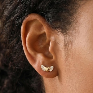May include: A close-up of a person's ear adorned with a pair of gold-colored stud earrings. Each earring features a wing-like design with small, clear gemstones. The earrings are set against the backdrop of the person's dark hair and skin.