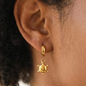 Può includere: Un orecchino a cerchio color oro con un piccolo ciondolo a forma di tartaruga lucida che pende dal fondo. L'orecchino è fissato al lobo dell'orecchio e presenta una finitura metallica liscia e riflettente sia sul cerchio che sul ciondolo.