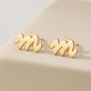May include: A pair of gold-toned stud earrings shaped like the lowercase letter m. Each earring features a smooth, polished surface with a cursive, flowing script design and a standard post backing for secure attachment to the earlobe.