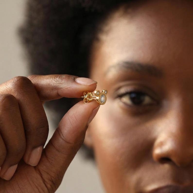 May include: A gold stud earring with a small, round, clear gemstone at the center. The earring is held between fingers, showcasing its delicate design and sparkling details. The earring has a halo of smaller gemstones around the central stone.