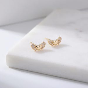 May include: A pair of gold-coloured stud earrings. Each earring features a heart shape at the centre, with small, clear stones forming wing-like accents on either side. The earrings are displayed on a white surface.