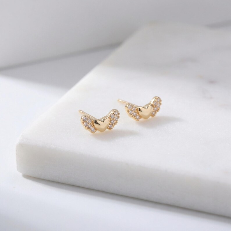 May include: A pair of gold-coloured stud earrings. Each earring features a heart shape at the centre, with small, clear stones forming wing-like accents on either side. The earrings are displayed on a white surface.
