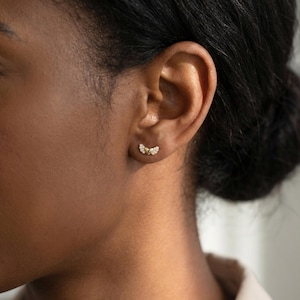 May include: A close-up of an ear displaying a pair of gold stud earrings. Each earring has a curved design, set with small, sparkling gemstones. The earrings are positioned against the skin, accentuating their intricate details.