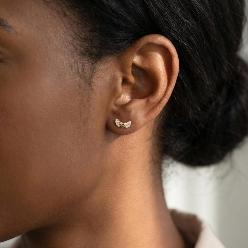 May include: A close-up of an ear displaying a pair of gold stud earrings. Each earring has a curved design, set with small, sparkling gemstones. The earrings are positioned against the skin, accentuating their intricate details.