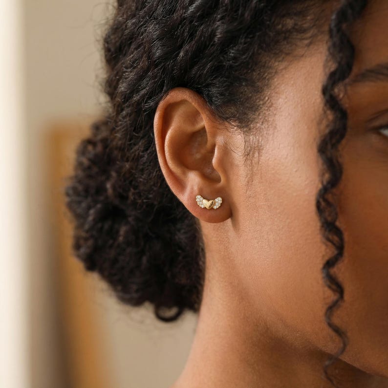May include: A close-up of a person's ear adorned with a pair of gold-colored stud earrings. Each earring features a wing-like design with small, clear gemstones. The earrings are set against the backdrop of the person's dark hair and skin.