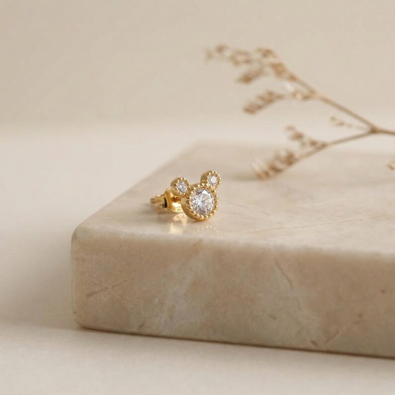 May include: A gold-colored stud earring in the shape of Mickey Mouse's head, with a large, round, clear gemstone in the center of the head and smaller gemstones for the ears. The earring is set on a light-colored surface.