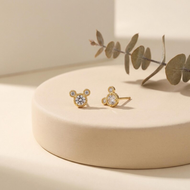 May include: A pair of gold-colored stud earrings in the shape of a mouse head. Each earring features a large, round, clear gemstone in the center, with smaller gemstones forming the ears. The earrings are displayed on a beige surface.