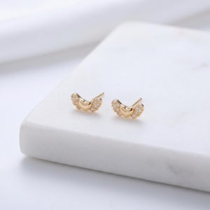 May include: A pair of gold stud earrings. Each earring has a heart-shaped centre with small, clear gemstones and wing-like details. The earrings are presented on a white surface, highlighting their delicate design and sparkle.
