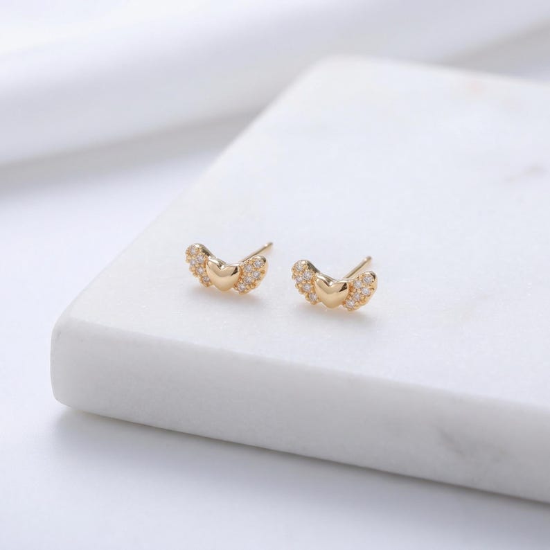 May include: A pair of gold stud earrings. Each earring has a heart-shaped centre with small, clear gemstones and wing-like details. The earrings are presented on a white surface, highlighting their delicate design and sparkle.