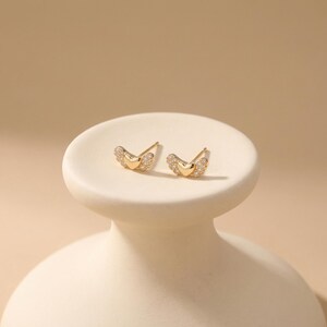 May include: A pair of gold-coloured stud earrings. Each earring has a heart-shaped centre and wing-like accents encrusted with small, clear gemstones. The earrings are displayed on a white, pedestal-style stand against a neutral background.