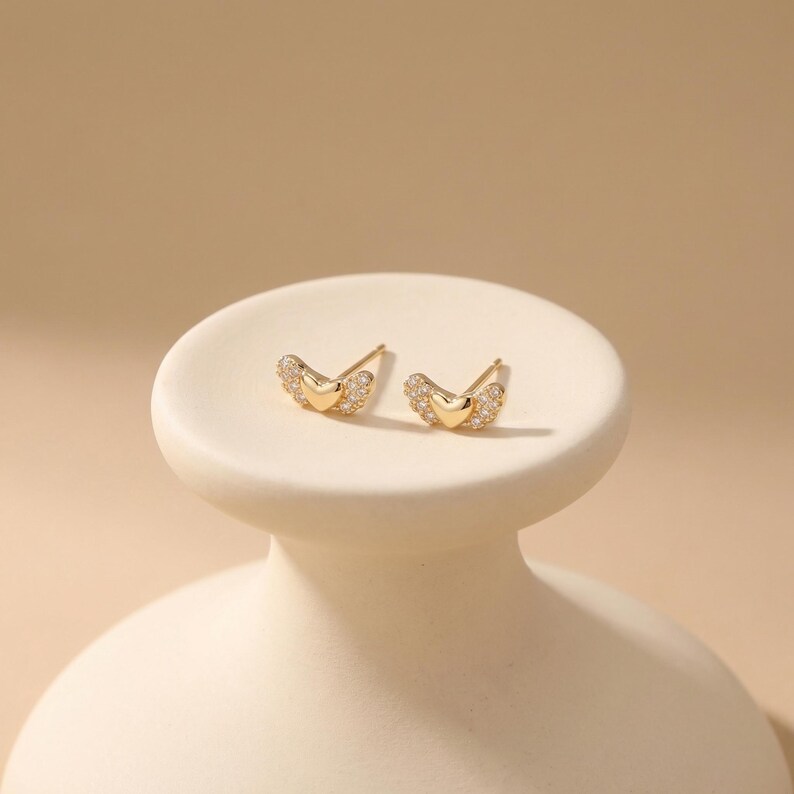 May include: A pair of gold-coloured stud earrings. Each earring has a heart-shaped centre and wing-like accents encrusted with small, clear gemstones. The earrings are displayed on a white, pedestal-style stand against a neutral background.
