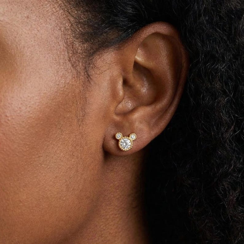 May include: Gold-toned stud earrings in a Mickey Mouse design. The center of the mouse's head features a round, clear gemstone surrounded by smaller clear stones. The ears are represented by two smaller, round clear stones.