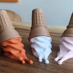 May include: Three whimsical ice cream cone sculptures with brown waffle cone tops and colorful, drippy "ice cream" in orange, white, and pink. The cones are arranged on a wooden surface, creating a playful and artistic display.