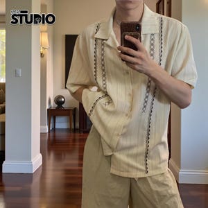 Könnte beinhalten: Cremefarbenes Kurzarmhemd und passende Shorts. Das Hemd hat vertikale Streifen und dekorative schwarze Nähte auf der Vorderseite und den Ärmeln. Das Hemd hat ein Knopfdesign und einen klassischen Kragen. Das Bild enthält den Text "Aldrix STUDIO".