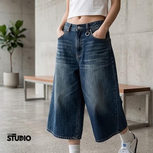 Pode incluir: Culotes de ganga azuis com lavagem desbotada. As calças de perna larga têm bolsos frontais e um detalhe de anel de metal na cintura. A modelo usa um top branco curto e ténis brancos. O logótipo "Aldrix Studio" é visível no canto inferior esquerdo.