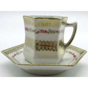 May include: A white porcelain teacup and saucer set with a hexagonal shape. The set features a delicate floral design with pink roses and gold trim.