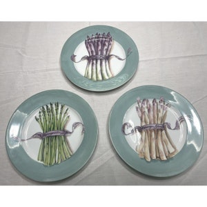May include: Three ceramic plates with a green rim and a white center. Each plate features a bunch of asparagus tied with a purple ribbon. The asparagus on the plates are purple, green, and white.