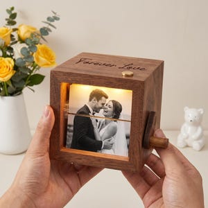 Puede incluir: Un cubo de fotos de madera con las palabras "Forever Love" grabadas en la parte superior. El cubo tiene un mecanismo giratorio para mostrar una foto de boda. El cubo está iluminado desde dentro y tiene un pequeño botón dorado en la parte superior.