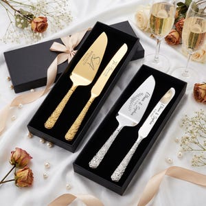 May include: Two cake serving sets in black boxes. One set is gold-toned with engraved text and floral handles. The other set is silver-toned with engraved text and floral handles. Champagne glasses and flowers are in the background.