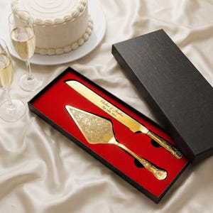 May include: A gold-toned cake knife and server set with intricate floral engravings on the server and the engraved text Chelsea & Jaylen Sep 24th, 2026 on the knife blade, presented in a black gift box with a vibrant red velvet interior lining.
