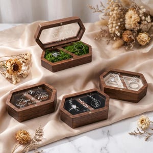 May include: Wooden hexagonal ring boxes with clear lids. The boxes are open, revealing rings nestled in moss. Some boxes have names and dates engraved on the lids. The boxes are on a cream-colored surface.