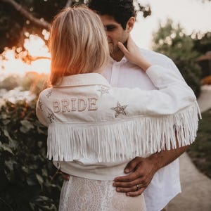 Puede incluir: Una chaqueta vaquera blanca con largos flecos en la espalda, adornada con la palabra BRIDE en letras nacaradas y varios parches brillantes en forma de estrella. La chaqueta se lleva sobre una prenda de encaje blanco con cuello.