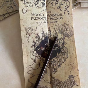 Puede incluir: Un mapa marrón estilo pergamino con el texto "MOONY, WORMTAIL, PADFOOT & PRONGS are proud to present" y "The Marauder's Map". Una varita marrón oscuro se coloca en el mapa. El mapa presenta una ilustración de castillo y remolinos decorativos.