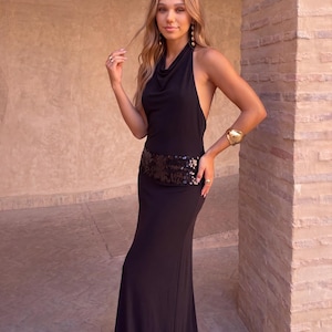 Puede incluir: Un vestido largo negro de cuello halter con escote tipo cowl y una cintura ancha adornada con lentejuelas negras. La prenda presenta un diseño de espalda descubierta y una silueta ajustada que llega hasta el suelo, en tela negra lisa.